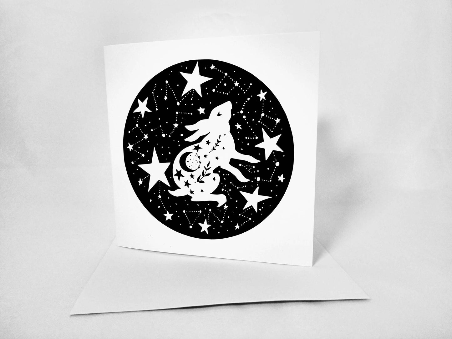 The Daring Hare Greeting Card - Cosmic Hare (5x5) features a white rabbit hare silhouette jumping within a black circular background filled with stars and constellations. The card, blank inside, is propped open on a white surface. The design is intricate and celestial-themed.