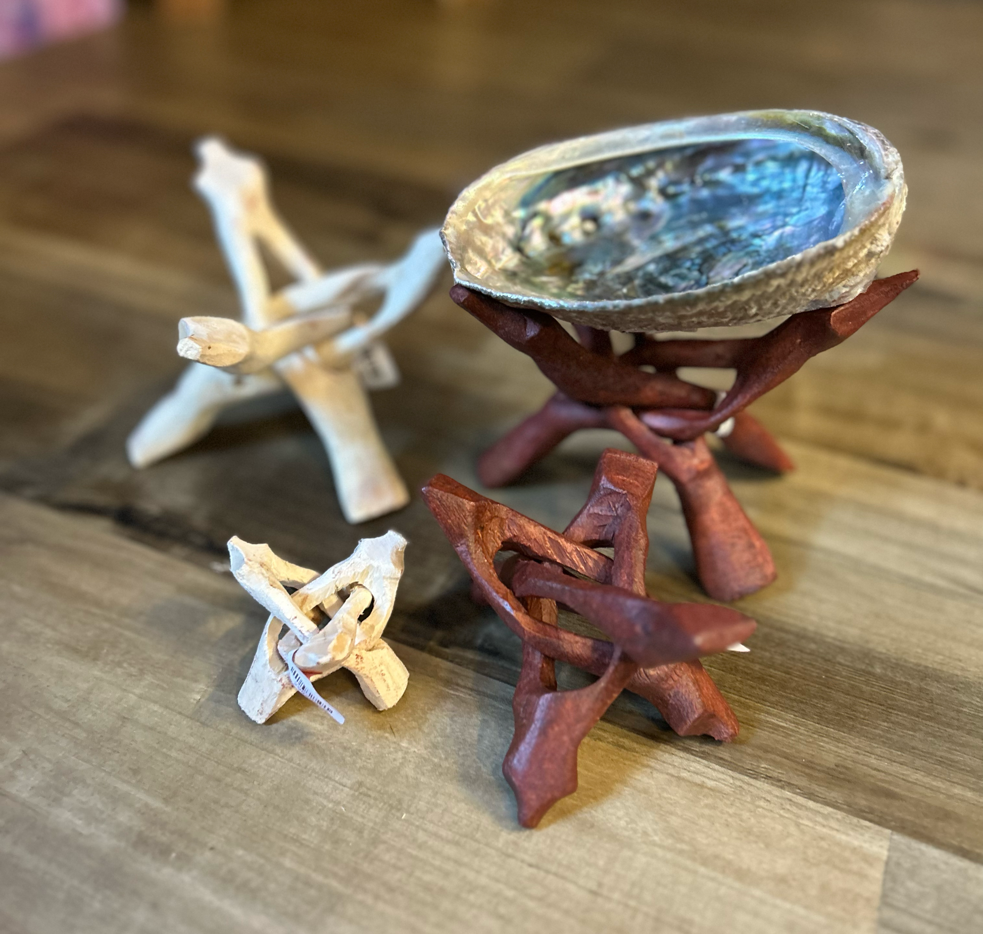 A wooden table hosts four wooden trivets, creating a serene sacred space. One trivet, placed at the top right corner, holds an abalone shell. The other three are empty and situated in front of it, with Atlanta Candle Carved Tripod Stand 6" varying in size and wood color: small white, medium brown, and large brown.