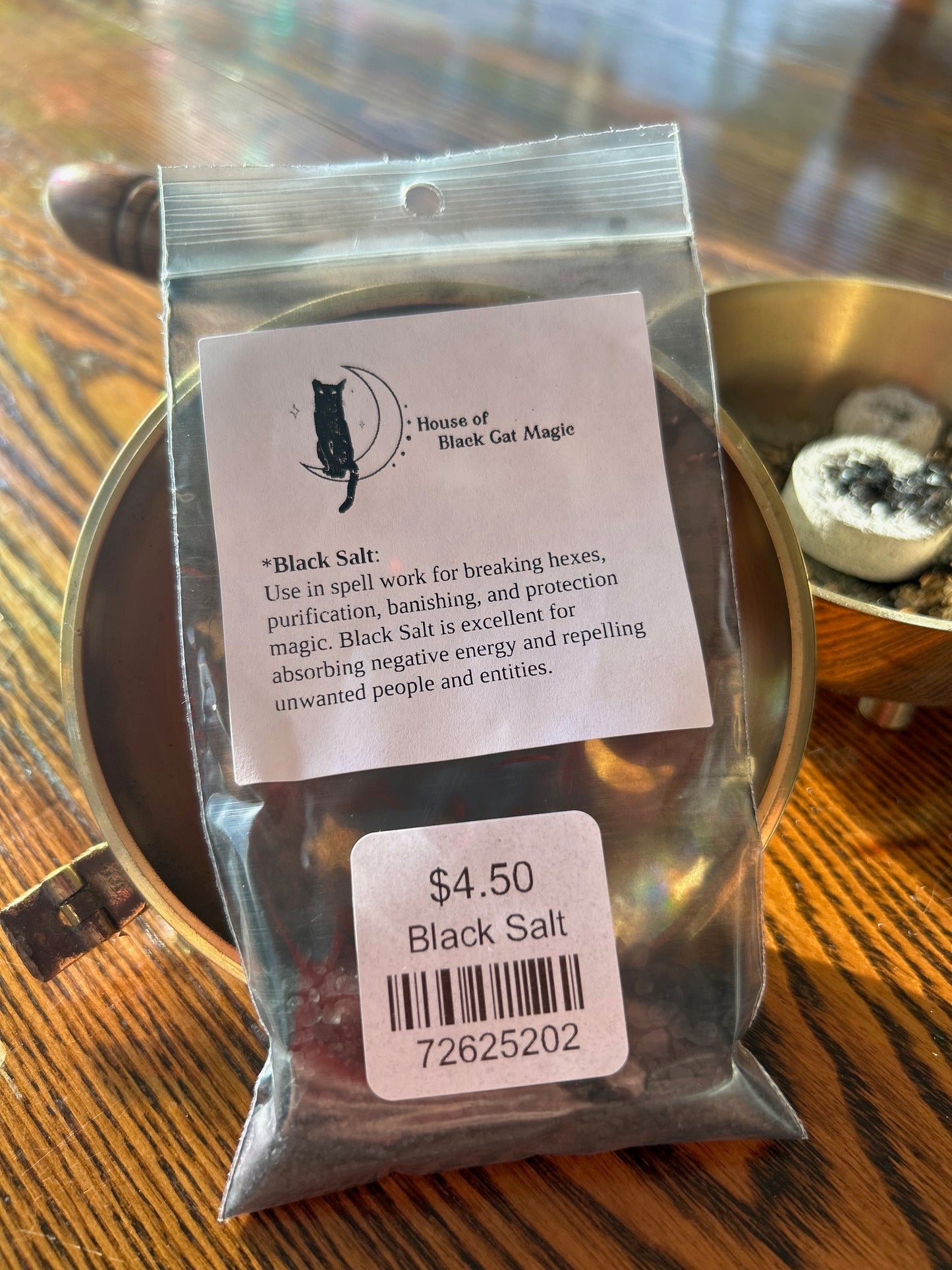 A bag of Black Salt from House of Black Cat Magic is displayed on a wooden surface. The label describes its use in breaking hexes, spellwork, purification, banishing negative energy, and protection magic. Priced at $4.50 with a barcode, a brass dish with charcoal discs is in the background.