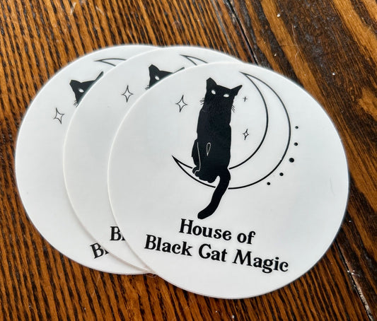 Three identical HBCM Circle Logo Stickers depict a feline sitting on a crescent moon with small stars scattered around. Below the image, the text reads "House of Black Cat Magic." Perfect for cat lovers, these stickers from House of Black Cat Magic are placed on a wooden surface.