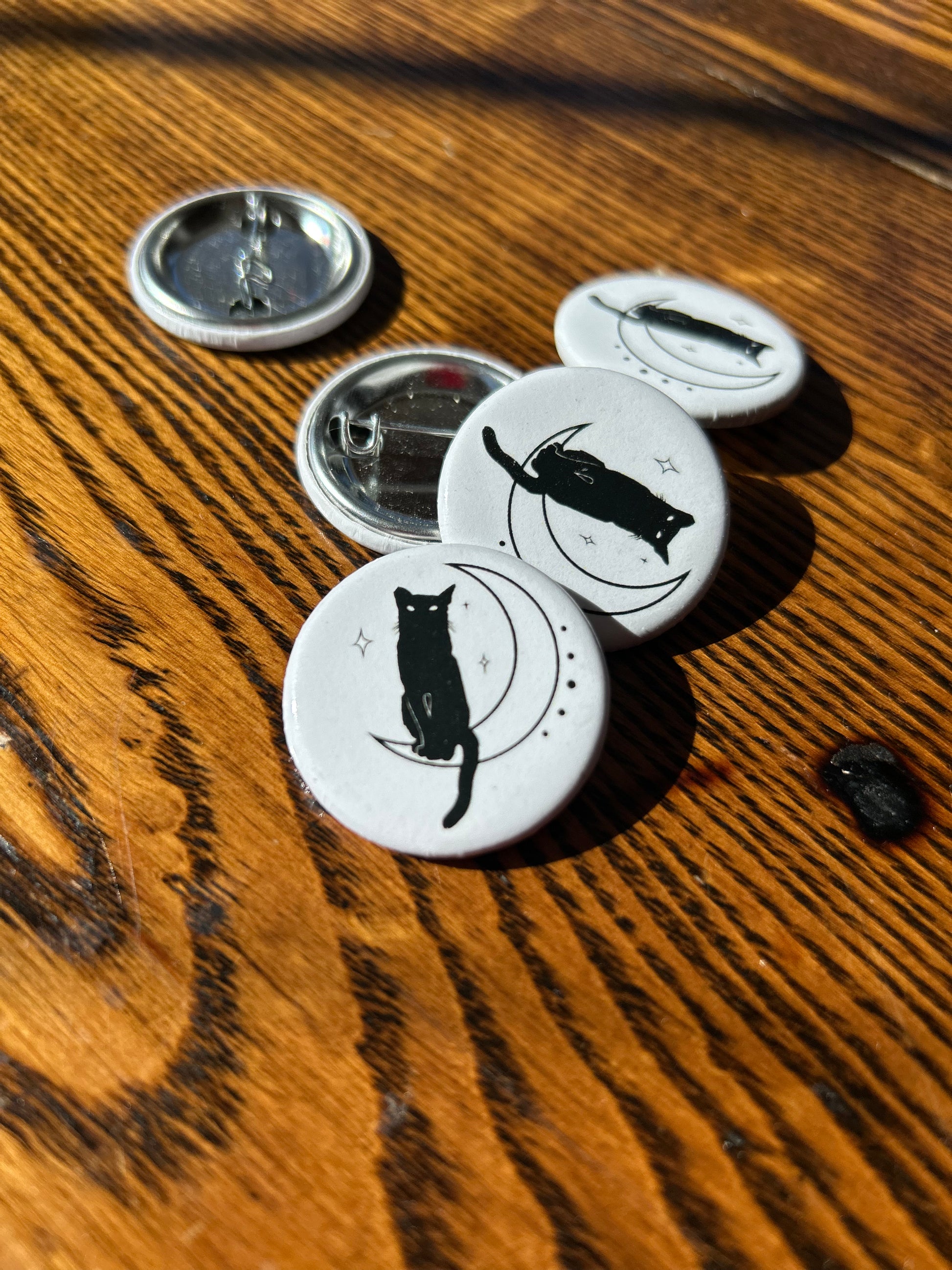 A group of HBCM Logo Buttons, embodying a witchy style and House of Black Cat Magic, are scattered on a wooden surface. One button depicts a feather. Some buttons are open, revealing the metal pin backs. The sunlight highlights the texture of the wooden surface.