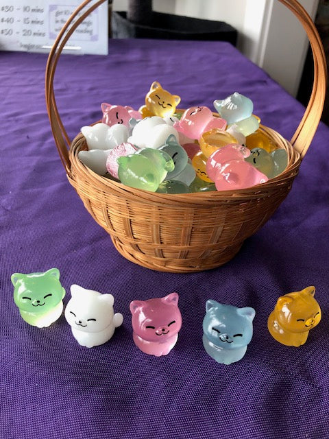 Alt text: A collection of one-inch tall cat figurines made of resin that glow in the dark after being exposed to sunlight. The figurines are arranged in rows on a white surface, showcasing their detailed features and smooth finish. Keywords: cat figurines, resin cat figurines, glow in the dark, one-inch tall figurines, cat collectibles, sunlight-activated glow, detailed cat figurines, small cat statues. Available in green, white, pink, blue, and orange.