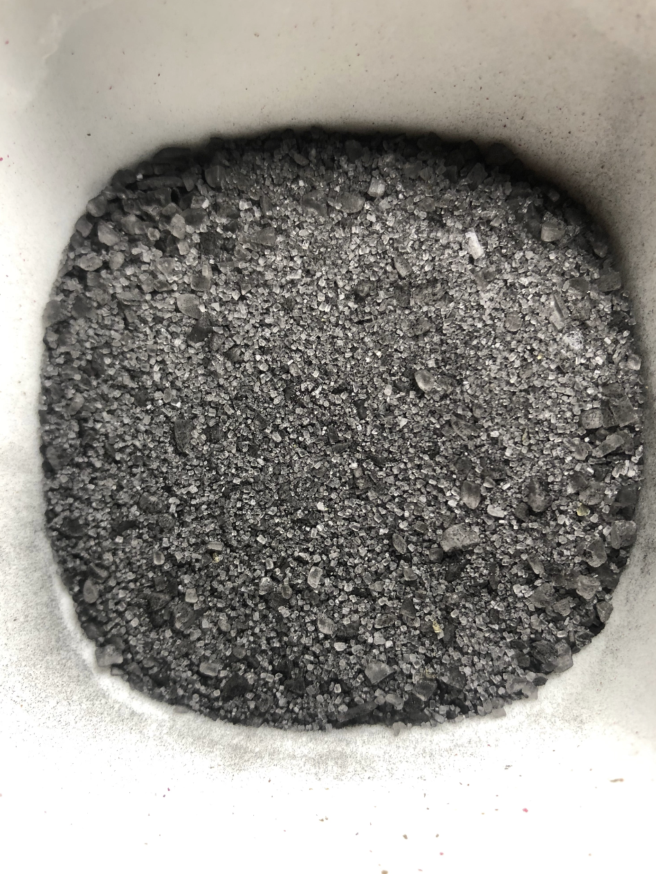 Close-up of a container filled with coarse, black crushed rock or gravel. The material, resembling Black Salt from House of Black Cat Magic often used in spellwork to ward off negative energy, appears granular and rough with varying sizes of dark, jagged particles. The sides of the container are partially visible, showing a white or grayish surface.